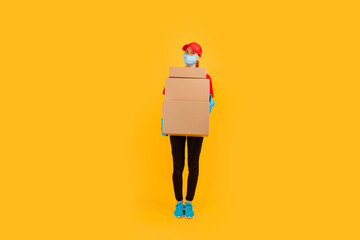 Full length, young female courier dressed in a Red cap and t-shirt, uniform, medical protective mask and gloves, holds cardboard boxes and shows a gesture of strength against a yellow background