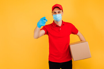 Fototapeta na wymiar male courier in a red uniform, medical mask and gloves holds a cardboard box and points to a copy of the space on a yellow background. Delivery service