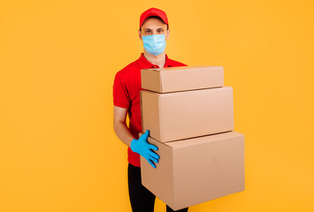 courier in a red uniform and a medical protective mask on his face holds cardboard boxes on an isolated yellow background. Online delivery, quarantine, coronavirus