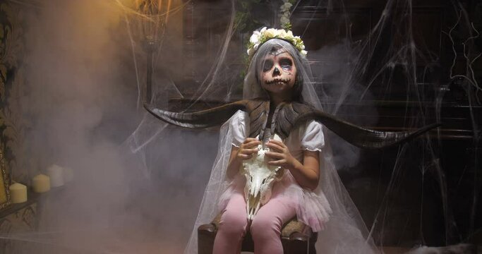 Portrait of girl dressed in Halloween Santa muerte costume  holding a goat scull in of old dark Gothic interior covered with cobwebs and mysterious fog and  changing lighting. 4K slow motion 50 fps