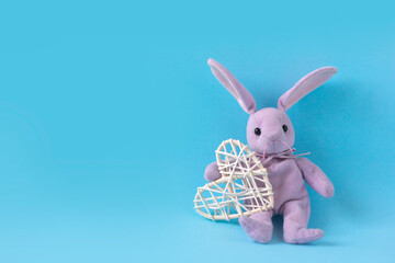 Pink plush hare sits on blue background and holds white heart. Valentine's day card