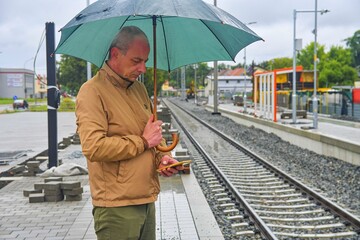 Portrait of a mature traveller texting on smartphone with an umbrella standing on the platform of a train station. Unfinished railway line. When the train arrives.. Funny concept of futile situation