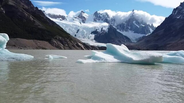 Drone fly over water along big glaciers in lake water called Laguna Torre against view of Cerro Torre peak in clouds and Fitz Roy. Los Glaciares National Park, Patagonia, Argentina