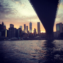 Going under the Brooklyn Bridge in a ferry, view of lower Manhattan skyline at sunset, Tall New York City skyscrapers late evening, dramatic colorful sky. Sun setting on Manhattan skyline.