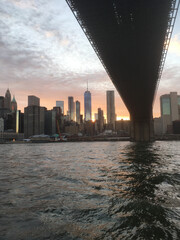 Going under the Brooklyn Bridge in a ferry on the East River, view of lower Manhattan skyline at sunset, Tall New York City skyscrapers late evening, dramatic colorful sky. Sun setting on Manhattan.