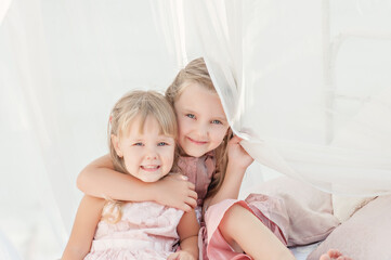 Two best blonde sisters in pink dresses are playing together. The girlfriends are hugging sitting on a white bed with chiffon canopies, decorated with wisteria. Strong friendship since childhood.