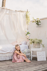 Obraz na płótnie Canvas Delicate little girl blonde with long hair in a beautiful pink powdery dress sits by a white bed with chiffon canopies, decorated with wisteria, flowers against the background of the sea. Design 