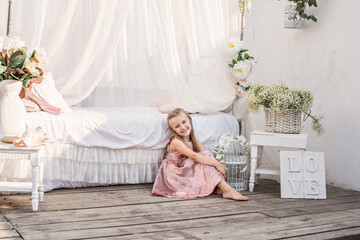 Obraz na płótnie Canvas Delicate little girl blonde with long hair in a beautiful pink powdery dress sits by a white bed with chiffon canopies, decorated with wisteria, flowers against the background of the sea. Design 
