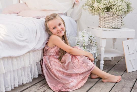 Delicate little girl blonde with long hair in a beautiful pink powdery dress sits by a white bed with chiffon canopies, decorated with wisteria, flowers against the background of the sea. Design 