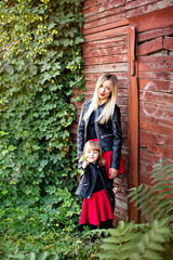 Happy beautiful mother and blonde daughter in the same family look, red skirts, black leather jackets, stand by a wooden brown barn, green ivy hops around. Mom hugs the girl, holds in her arms. Day 