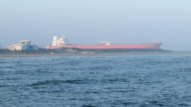 Ocean sailing oil tanker ship approaches the waters of the Gulf of Mexico in Port Aransas, Texas on a hazy evening, with waves and beach and copy space, in slow motion clip.