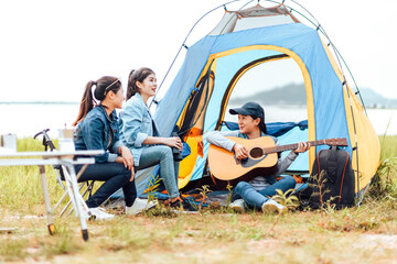 A group of friends camping happily near the lake on a holiday. A woman plays the guitar for a friend to listen.