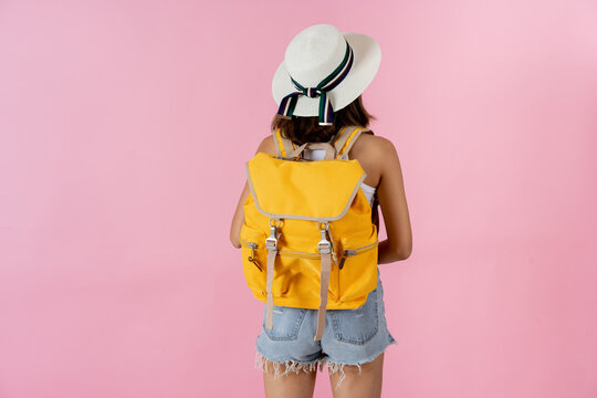 Rear View Of Young Woman Going On Vacation Wearing Sunhat And Carrying Yellow Backpack Against Plain Pink Background