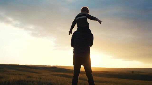 happy family. son sits on his neck teamwork at father shows hands lifestyle to the side plays at the pilot depicts an airplane silhouette at sunset. happy family concept childhood man dad with little