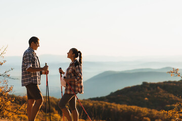 Young pregnant woman with boyfriend nordic walking outdoors with trekking poles. Active and healthy lifestyle in maternity time. Beautiful autumn mountain view. - 364635355