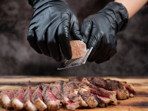 hand in black glove scratch Himalayan stone salt to sliced beef steak on wooden cutting board.