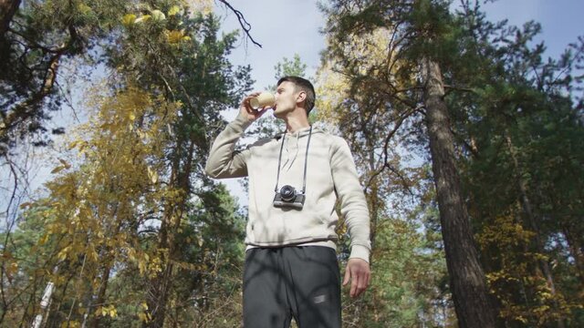 4K portrait of caucasian man in sportswear with camera hanging from his neck drinking from cardboard cup and looking around at autumn forest. Low angle 360 degree tracking arc shot.