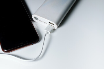 A white power bank with USB ports and power button. A cable is attached to it and a smart phone is being charged