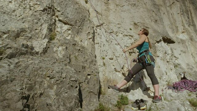 SLOW MOTION, CLOSE UP: Female climber belays her friend climbing up a difficult stone wall as her cute puppy calmly sits behind her and observes the climbers in action. Woman belaying a rock climber.