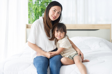 Lovely Asian mother and little daughter hug together, smile and feeling happy on the bed in a cozy house, look at camera, family concept