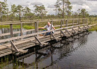 Fototapeta na wymiar the summer swamp. a man in a white shirt sits on a wooden bridge. bog pond. bog background and vegetation