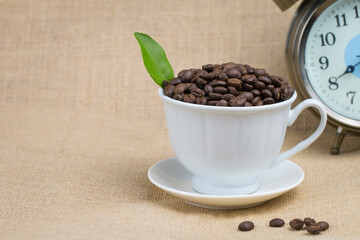 Roasted coffee beans In a white coffee cup and with an alarm clock and the bottom surface is a brown fabric and a few coffee beans scattered on the floor and the image is empty and has green leaves