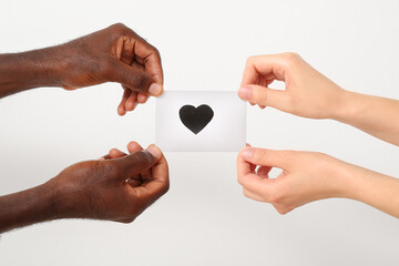 Hands of Caucasian woman and African-American man holding paper sheet with drawn heart on light...