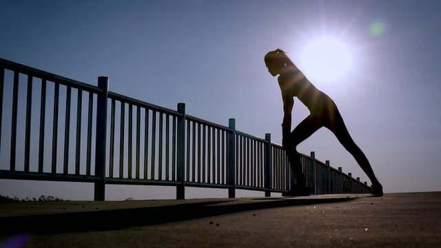 silhouette of sporty woman preforming stretching her muscle, worming up before running workout at outdoors street