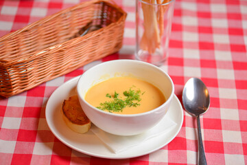 Pumpkin soup in a white bowl. Wooden table, red plaid tablecloth, blurred background. Dinner in restaurant.