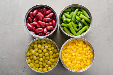 Flat lay view at canned kidney beans, green beans, peas and corn in opened tin cans on kitchen table. Non-perishable foods background
