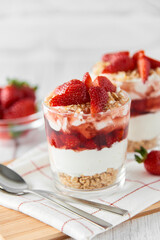 Homemade layered dessert with fresh strawberries, cream cheese or yogurt, granola and strawberry jam in glasses on white wood background. Healthy organic breakfast or snack concept. Selective focus.