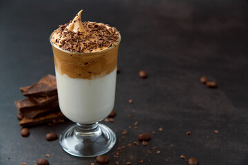 Glass of trendy fluffy creamy dalgona or whipped instant coffee with milk, coffee beans and chocolate on the dark background. New popular food and drink trend concept. Copy space.