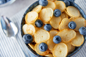 Blue Bowl of mini pancake cereal with blueberries, maple syrup and milk on concrete background. Tiny pancakes, new food trend concept. Cute healthy breakfast or snack. Recipe, menu.