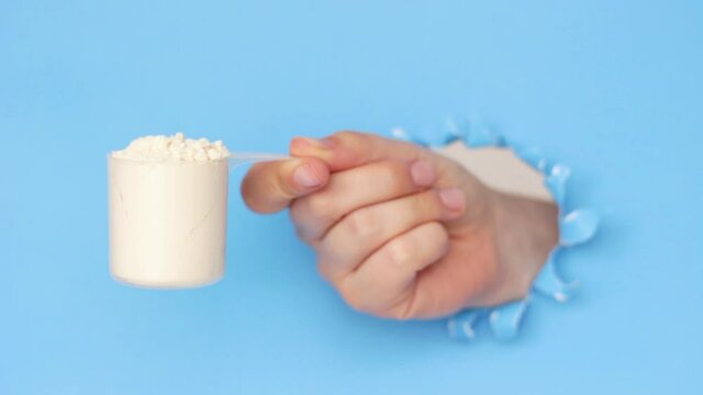 The woman's hand holds the protein in measuring cup on a blue background