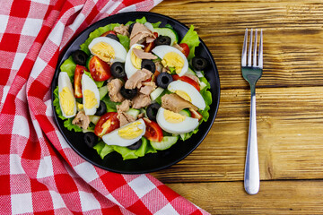 Tasty tuna salad with lettuce, black olives, eggs and fresh vegetables on wooden table. Top view