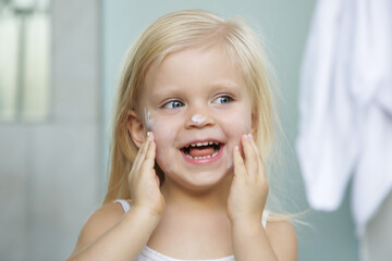 Adorable blonde caucasian toddler girl applying cream on her face 