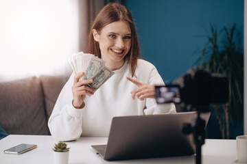 Smiling female blogger with brown hair sitting at table with dollars banknotes while recording...