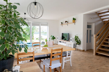 Dining room with table and chairs in white bright interior with wooden floor and stairs. Bright white interior with green plants and tv. Stylish and scandinavian decor.