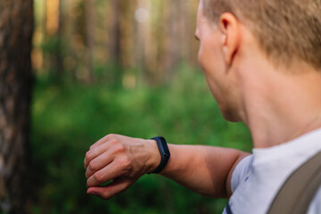 A young blond man looks at a watch or fitness bracelet, leads an active lifestyle and walks in the woods