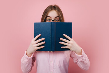 Emotional blonde female in eyeglasses hiding her face behind opened book over pink background....