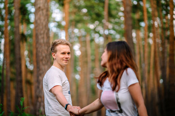 Obraz premium A young active couple of young people hold each other's hands while taking a walk through a pine forest, one of the lovers blurred