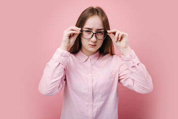 Beautiful blonde girl in pink shirt holding hands on eyeglasses and looking at camera with suspicious glance. isolated over studio background. Concept of real human emotion.