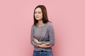 Portrait of sad young girl wincing in pain and clutching belly beacuse of strong stomach ache. Isolated over pink studio background. Concept of spasms.