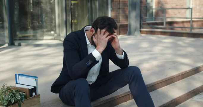 Depressed man holding head with hands while sitting near box with stuff on office building stairs . Male worker looking frustrated after losing job in crisis. Concept of unemployment