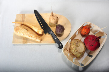 Root vegetables in basket and on wooden cutting board with knife.  High angle view with white...