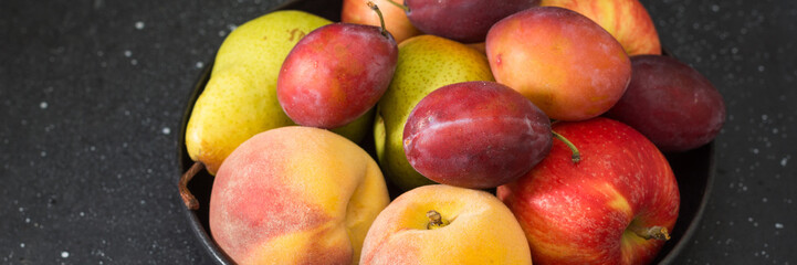 Various fruits in a black ceramic plate on a black kitchen table. Apples, plums and peaches on a plate. Banner with space for text