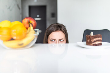 Hard choice. Yound sporty woman is choosing between fruits and sweets while standing on light kitchen. Junk food or healthy food.