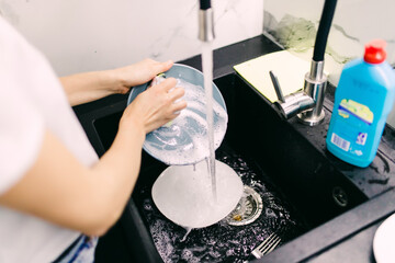 Hands washing dishes with running water from faucet in sink
