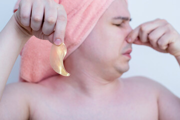 A young discontented frowning man puts gold bandages under his eyes, removes wrinkles and dark circles, and takes care of the delicate skin under the eyes. A man doesn't like makeup.