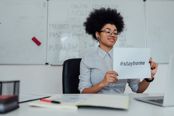 Young happy afro american female teacher showing paper with text STAY HOME at webcam while sitting at her workplace and teaching English online